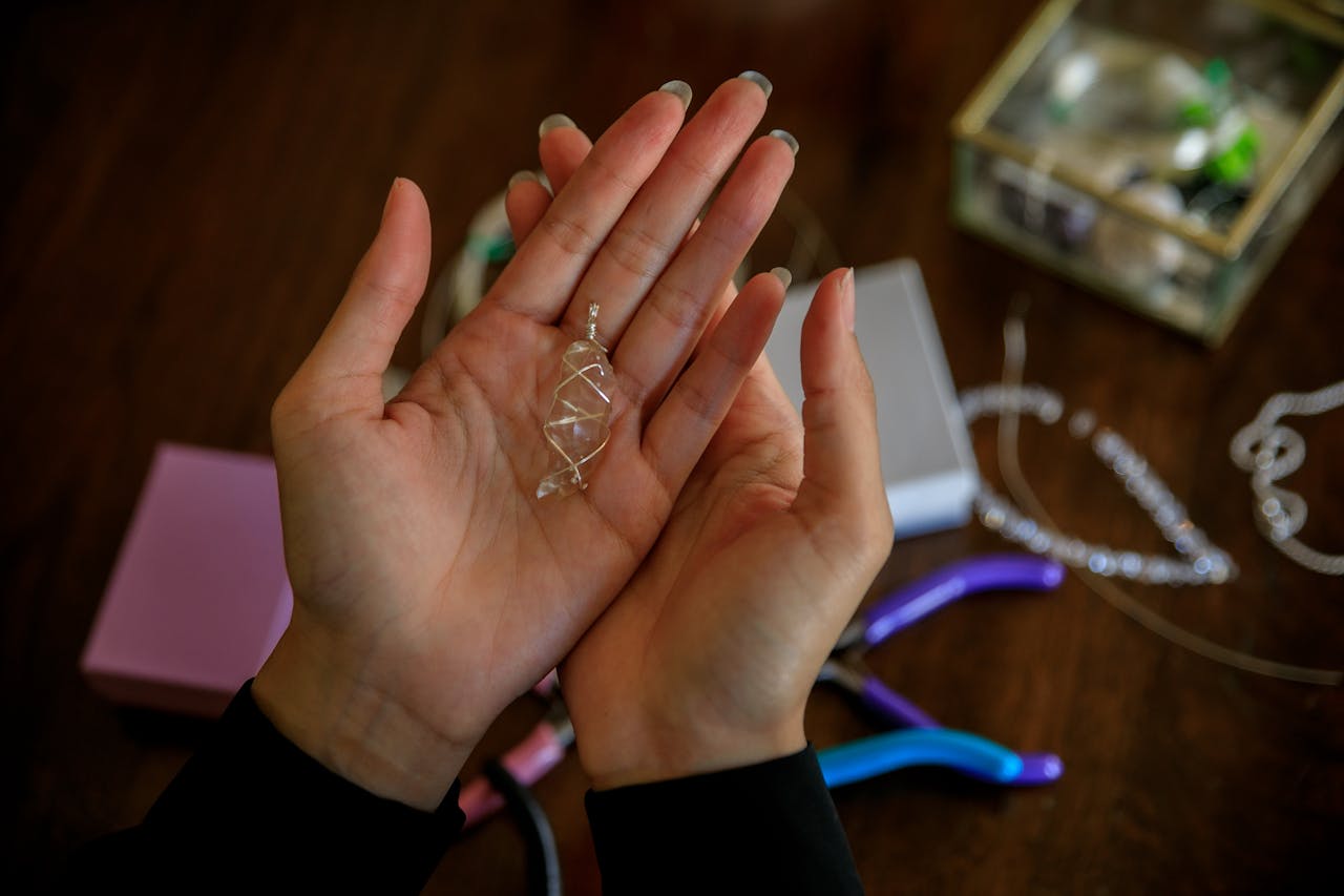about-us-02 Close-up of hands presenting a handmade silver wire pendant with jewelry tools on a wooden table.